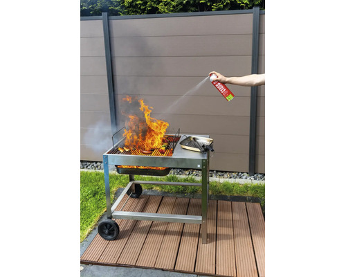 Person löscht Flammen auf einem Holzkohlegrill mit einer Feuerlöschspraydose im Garten vor einem Sichtschutzzaun.