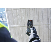Hand in Arbeitshandschuh hält ein digitales Bosch Laser Entfernungsmessgerät mit Display vor einem Bauzaun.