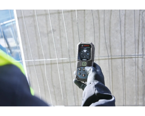 Hand in Arbeitshandschuh hält ein digitales Bosch Laser Entfernungsmessgerät mit Display vor einem Bauzaun.