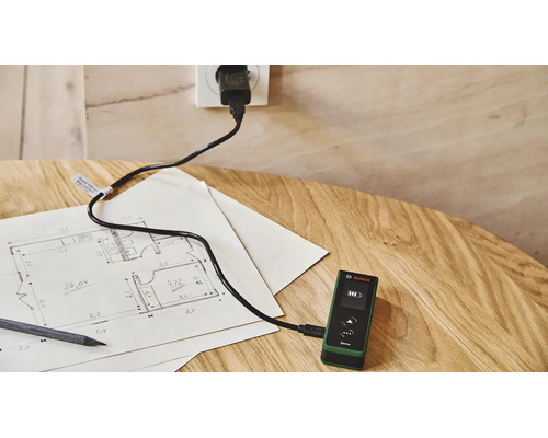 Bosch Laser Entfernungsmesser Zamo wird mit einem USB Kabel an einer Steckdose auf einem Holztisch mit einem Grundrissplan geladen