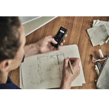 Eine Person verwendet einen Bosch Laser Entfernungsmesser beim Zeichnen eines Raumplans auf Papier an einem Holztisch.