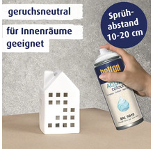Eine Hand hält eine Sprühdose Belton Aqua Colours Lack, um ein kleines Haus aus Keramik zu besprühen, mit Hinweisen zu Geruchsneutralität und Abstand.