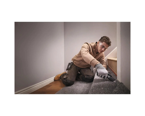 Handwerker mit Schutzbrille und Handschuhen beim Zuschneiden von Teppichboden auf einer Treppe mit einem Teppichmesser.