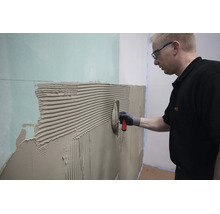 Handwerker beim Auftragen von Fliesenkleber mit einer Zahnkelle auf eine Gipskartonwand