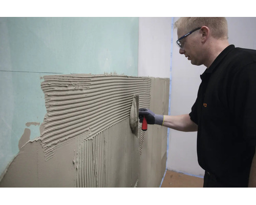 Handwerker beim Auftragen von Fliesenkleber mit einer Zahnkelle auf eine Gipskartonwand