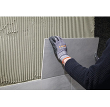 Handwerker mit Arbeitshandschuhen bringt Fliese auf mit Fliesenkleber gestrichener Wand an