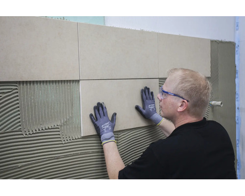 Handwerker mit Schutzbrille und Arbeitshandschuhen bringt Fliesen auf einer mit Fliesenkleber bestrichenen Wand an.
