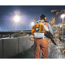 Ein Arbeiter mit Stihl Laubsauger auf dem Rücken reinigt bei Nacht einen Gehweg von Herbstlaub unter einer Straßenlaterne.