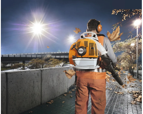 Ein Arbeiter mit Stihl Laubsauger auf dem Rücken reinigt bei Nacht einen Gehweg von Herbstlaub unter einer Straßenlaterne.