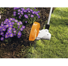 Ein elektrischer Rasentrimmer schneidet die Kante zwischen dem Rasen und einem Beet mit violetten Blumen.
