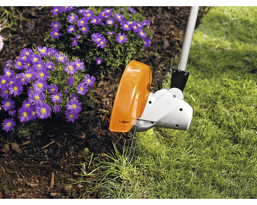 Ein elektrischer Rasentrimmer schneidet die Kante zwischen dem Rasen und einem Beet mit violetten Blumen.