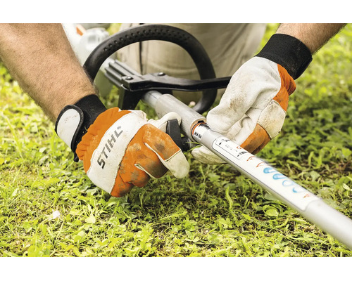 Eine Person montiert ein Stihl KombiWerkzeug an einen Stihl KombiMotor Schaft mit Handschuhen in einer Gartenumgebung.