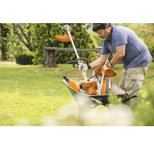 Ein Gärtner legt ein Stihl Kombigerät und einen Freischneideraufsatz in eine Schubkarre auf einer Wiese.