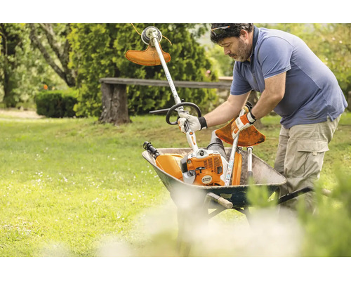 Ein Gärtner legt ein Stihl Kombigerät und einen Freischneideraufsatz in eine Schubkarre auf einer Wiese.