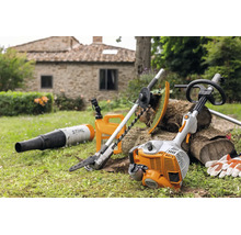 Stihl Kombimotor mit verschiedenen Aufsätzen wie Heckenschere und Laubbläser im Garten vor einem Steinhaus.