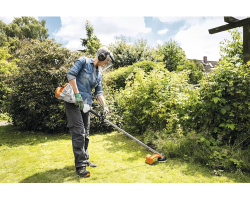 Eine Person benutzt einen Stihl Motorsensen Freischneider zur Rasenpflege in einem Garten mit schützender Arbeitskleidung und Gesichtsschutz.