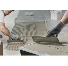 Handwerker beim Auftragen von Fliesenkleber mit einer Zahnkelle auf einem Boden für die Verlegung von Fliesen.