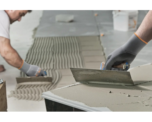 Handwerker beim Auftragen von Fliesenkleber mit einer Zahnkelle auf einem Boden für die Verlegung von Fliesen.