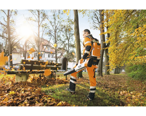 Ein Arbeiter verwendet einen Stihl Akku Laubbläser mit rückentragbarem Akku, um herbstliches Laub in einer Parkanlage zusammenzublasen.