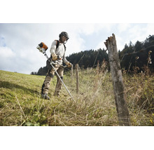 Eine Person benutzt eine Stihl Motorsense bei der Arbeit auf einer Wiese entlang eines Zauns mit persönlicher Schutzausrüstung.