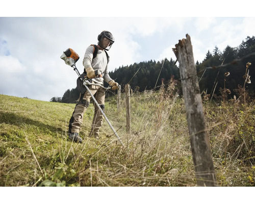 Eine Person benutzt eine Stihl Motorsense bei der Arbeit auf einer Wiese entlang eines Zauns mit persönlicher Schutzausrüstung.