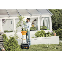 Ein Mann nutzt einen Stihl Elektro Häcksler GHE 105 im Garten vor einem Gewächshaus, um Zweige und Äste zu zerkleinern.
