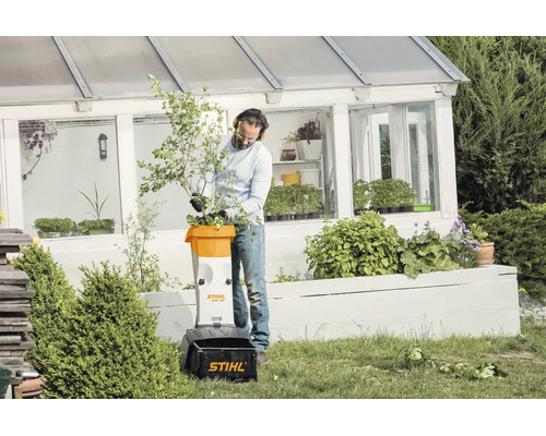 Ein Mann nutzt einen Stihl Elektro Häcksler GHE 105 im Garten vor einem Gewächshaus, um Zweige und Äste zu zerkleinern.
