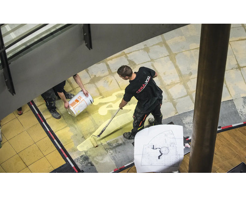 Handwerker beim Auftragen von Grundierung mit einer Farbrolle auf einem gefliesten Bodenbelag in einem Innenbereich.