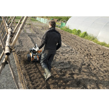 Eine Person nutzt eine Gartenfräse auf einem Ackerfeld in einem Gemüsegarten mit Gewächshaus.