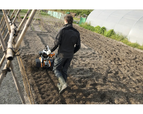 Eine Person nutzt eine Gartenfräse auf einem Ackerfeld in einem Gemüsegarten mit Gewächshaus.