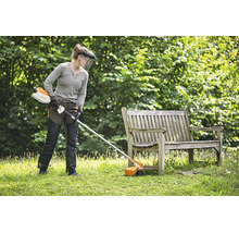 Person mit Schutzausrüstung nutzt einen Stihl Akku Motorsense zum Rasenmähen in einem Garten neben einer Holzbank.