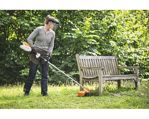 Person mit Schutzausrüstung nutzt einen Stihl Akku Motorsense zum Rasenmähen in einem Garten neben einer Holzbank.