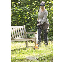 Eine Person mit Schutzkleidung verwendet einen Stihl Rasentrimmer, um Gras entlang einer hölzernen Gartenbank in einem Garten zu schneiden.