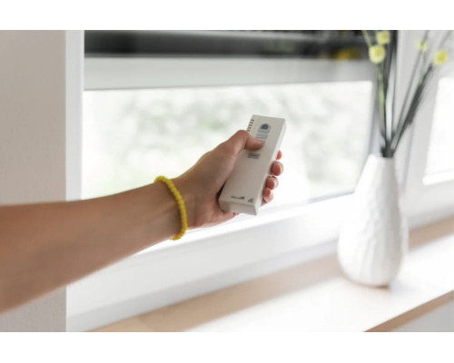 Eine Hand hält eine Fernbedienung zur Steuerung von Jalousien oder Rolläden vor einem Fenster.
