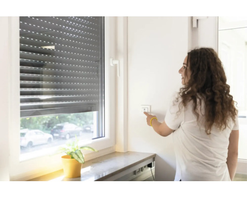 Eine Frau bedient einen elektrischen Rollladen an einem Fenster mit einem Wandschalter im Innenbereich.