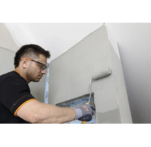 Handwerker mit Schutzbrille und Handschuhen trägt mit einer Farbrolle Grundierung auf eine Wand im Innenraum auf.