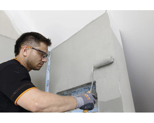 Handwerker mit Schutzbrille und Handschuhen trägt mit einer Farbrolle Grundierung auf eine Wand im Innenraum auf.