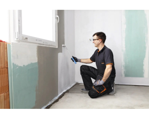 Handwerker streicht mit einer Farbrolle eine Innenwand in einem Raum unter einem Fenster.