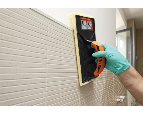 Hand mit Handschuh reinigt Fliesen an der Wand mit einem Fugengummi oder Fliesenwaschbrett