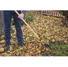Eine Person verwendet einen Laubrechen mit Holzstiel, um Herbstlaub im Garten zusammenzurechen.