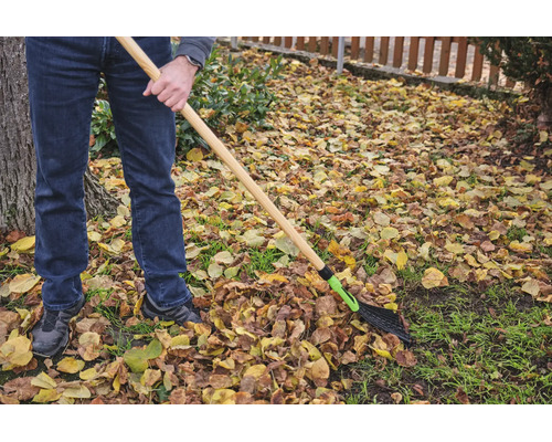 Eine Person verwendet einen Laubrechen mit Holzstiel, um Herbstlaub im Garten zusammenzurechen.