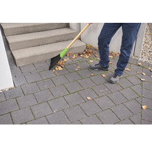Person reinigt eine gepflasterte Terrasse vor einer Treppe mit einem Straßenbesen mit Holzstiel