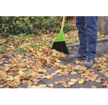 Eine Person kehrt mit einem Straßenbesen Herbstlaub auf einem asphaltierten Weg zusammen.
