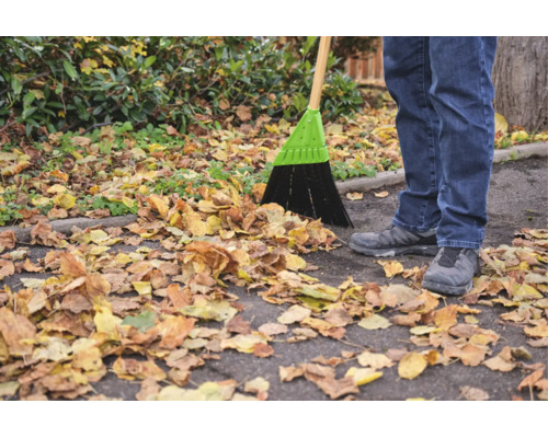 Eine Person kehrt mit einem Straßenbesen Herbstlaub auf einem asphaltierten Weg zusammen.