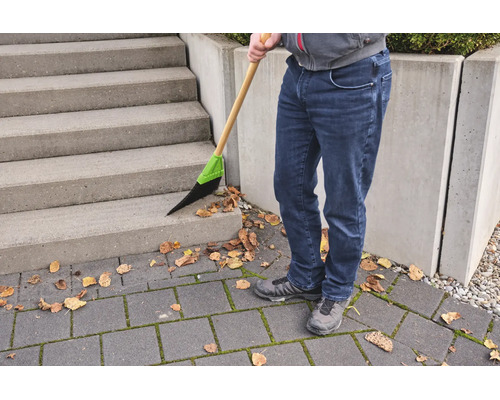 Person kehrt mit einem Besen mit Holzstiel und Kunststoffborsten trockenes Laub von einer Betontreppe und einem gepflasterten Boden.