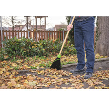 Eine Person benutzt einen Gartenbesen mit Holzstiel und Kunststoffborsten, um Herbstlaub von einem asphaltierten Weg in einem Garten zu kehren.