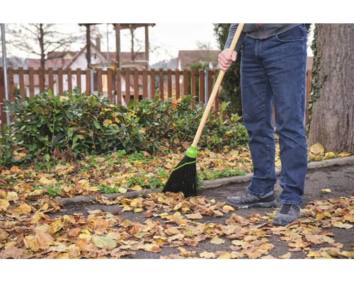 Eine Person benutzt einen Gartenbesen mit Holzstiel und Kunststoffborsten, um Herbstlaub von einem asphaltierten Weg in einem Garten zu kehren.
