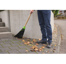 Person verwendet einen Straßenbesen mit Holzstiel, um herbstliches Laub von einem gepflasterten Gehweg zu kehren.