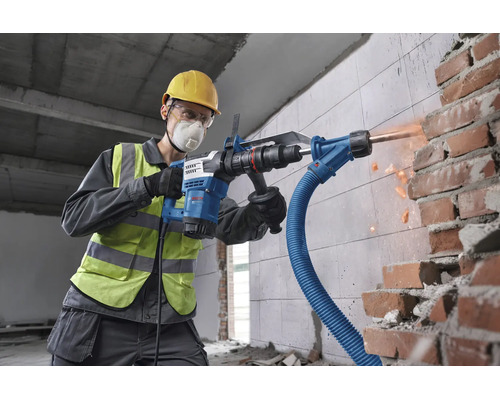 Handwerker mit Schutzausrüstung verwendet einen Bosch Bohrhammer mit Staubabsaugung beim Meißeln in einer Ziegelwand auf einer Baustelle.