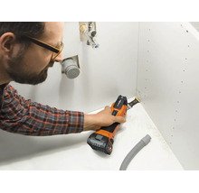 Ein Mann verwendet ein Akku Multitool um einen Ausschnitt in einer weißen Schrankplatte für Wasseranschlüsse zu sägen.